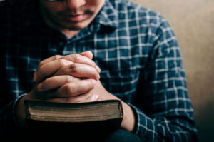 man praying with hands folded in on a bible